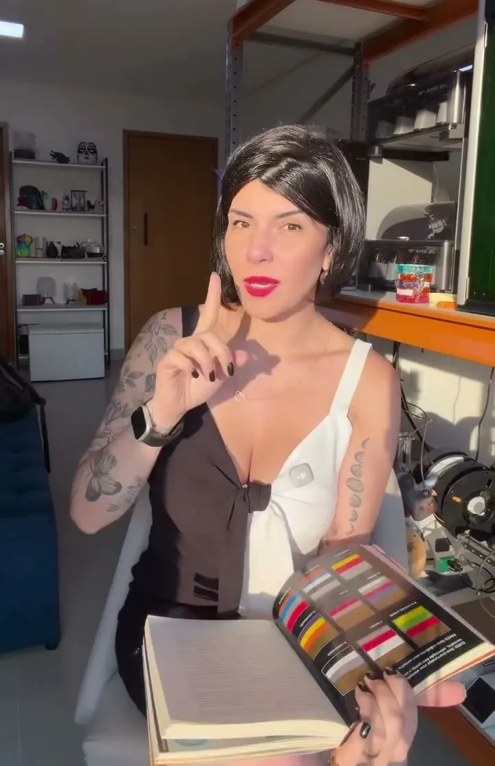 Carol Cardoso em pé no estúdio dela, segurando um mostruário de cores de filamento, com prateleiras de peças impressas ao fundo