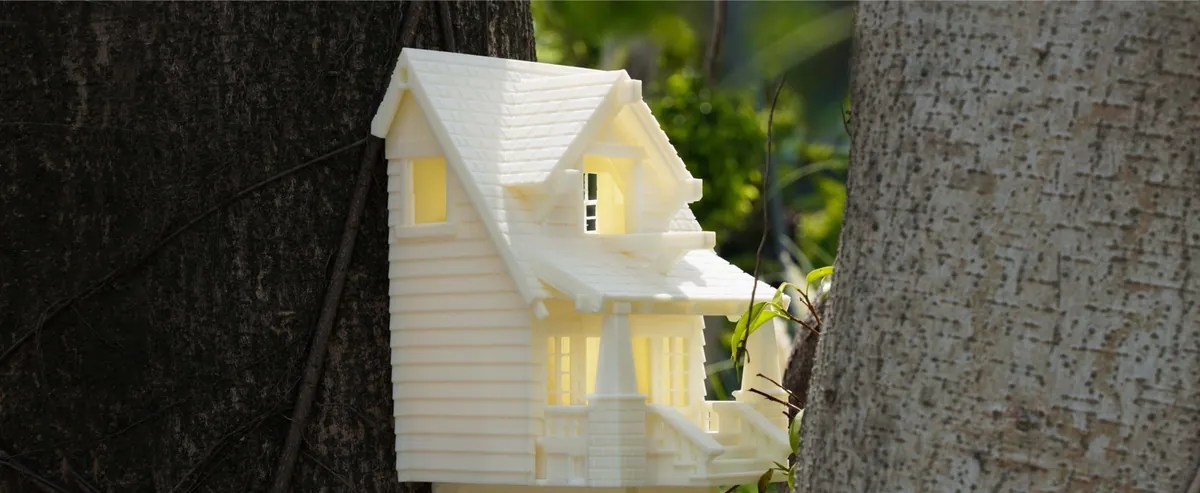 Vaso impresso em 3D em forma de casinha branca, com telhado, janelas e varanda, preso entre dois troncos de árvore ao ar livre