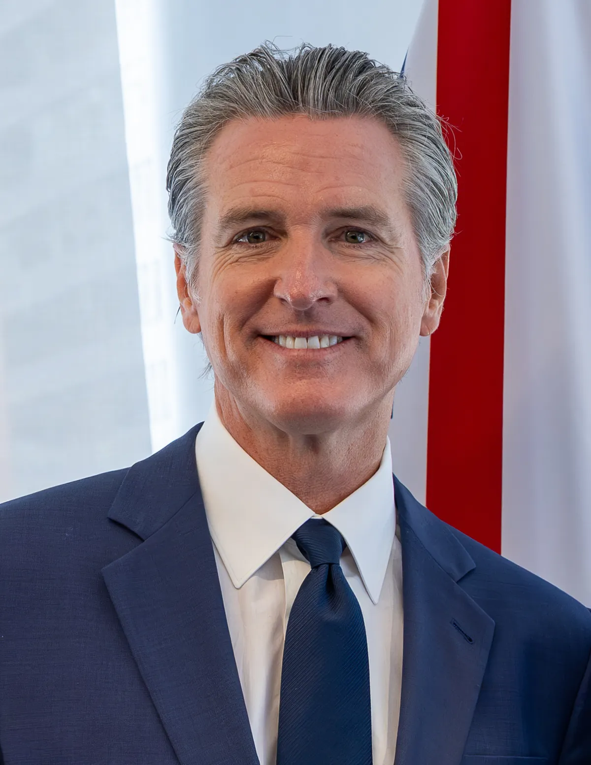 Retrato do governador da Califórnia, Gavin Newsom, de terno azul e gravata, sorrindo, com uma bandeira vermelha e branca desfocada ao fundo