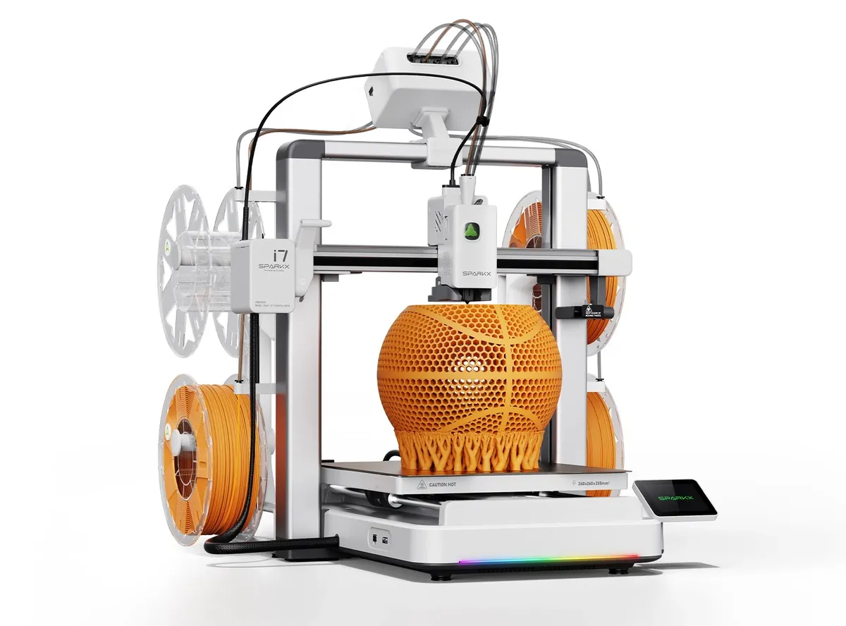 Impressora SPARKX i7 branca imprimindo uma bola de basquete vazada em filamento laranja, com bobinas laranja nas laterais e o kit CFS nano no topo