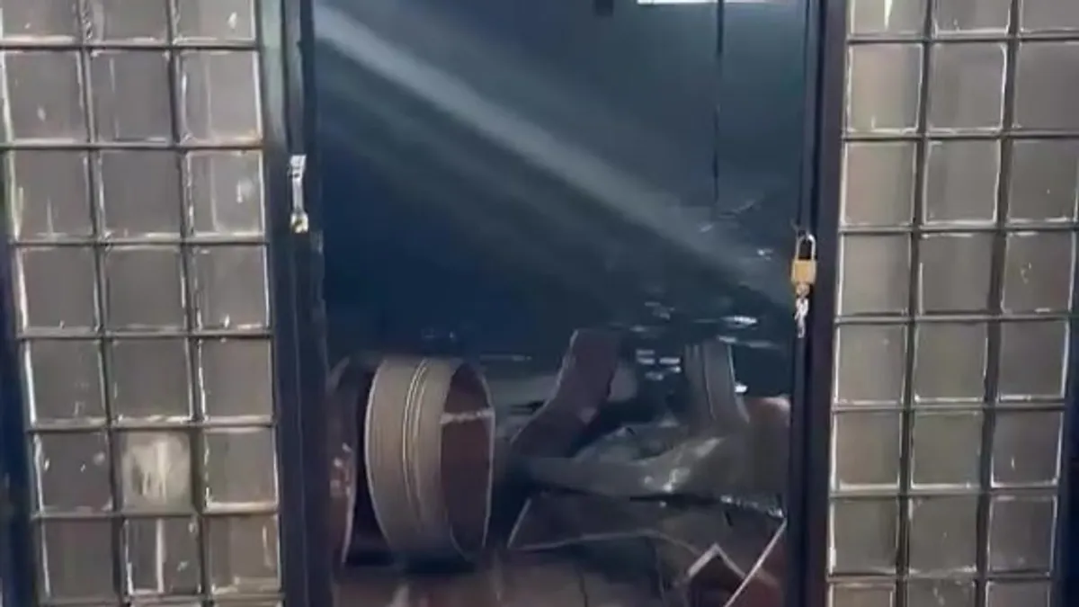 Entrada do galpão incendiado, com a porta aberta, luz atravessando a fumaça e destroços no chão