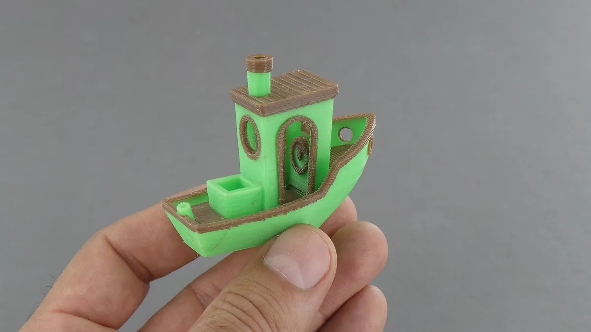 Mão segurando um 3DBenchy, o barquinho de teste da impressão 3D, impresso em verde com detalhes em marrom sobre fundo cinza
