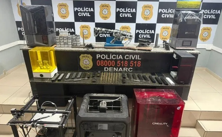 Impressoras 3D de resina e filamento, arma e carregadores apreendidos expostos na sede do Denarc, com painel da Polícia Civil ao fundo