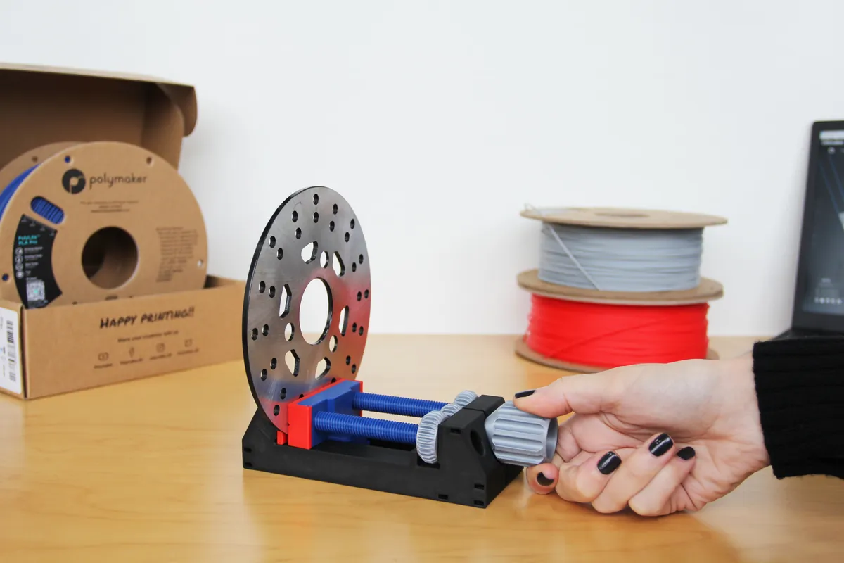 Rolo de filamento Polymaker PolyLite PLA Pro na caixa de papelão, ao lado de uma morsa impressa em 3D que segura um disco de freio e de dois carretéis de filamento cinza e vermelho