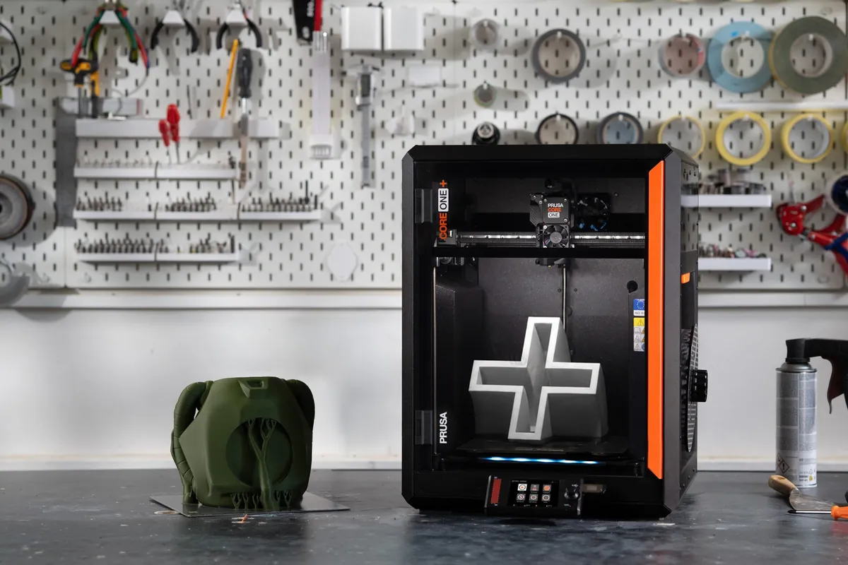 Impressora Prusa Core One+ preta, com detalhe laranja na lateral e uma peça branca em forma de cruz sobre a mesa, numa bancada de oficina com ferramentas penduradas ao fundo