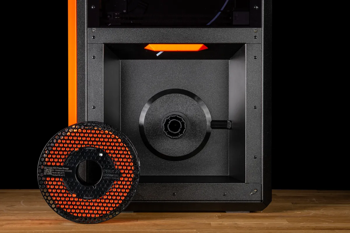 Compartimento traseiro de uma Prusa Core One com o suporte de carretel redondo e preto do kit OpenPrintTag instalado e, à frente, um carretel vazado de Prusament ASA laranja apoiado numa bancada de madeira