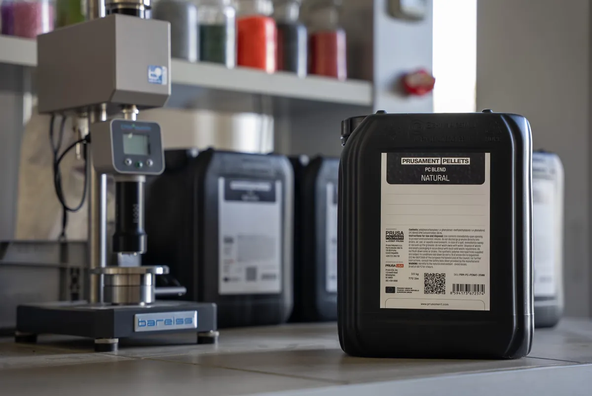 Galão preto de 5 litros com o rótulo Prusament Pellets PC Blend Natural sobre uma bancada de laboratório, ao lado de um durômetro e de outros galões