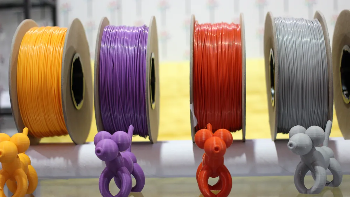 Rolos de filamento de varias cores numa prateleira, com uma peca impressa em 3D na frente de cada rolo