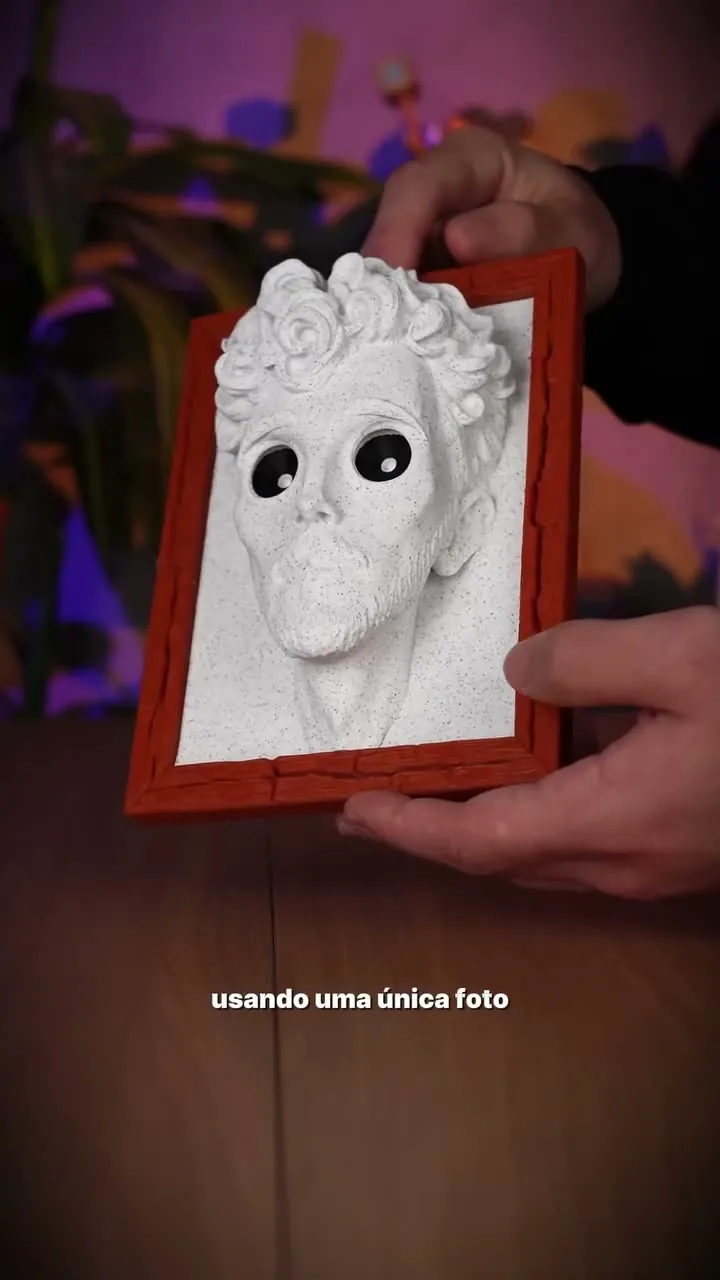 Mãos seguram um retrato impresso em 3D dentro de uma moldura vermelha, com os olhos vazados que criam o efeito de ilusão de ótica